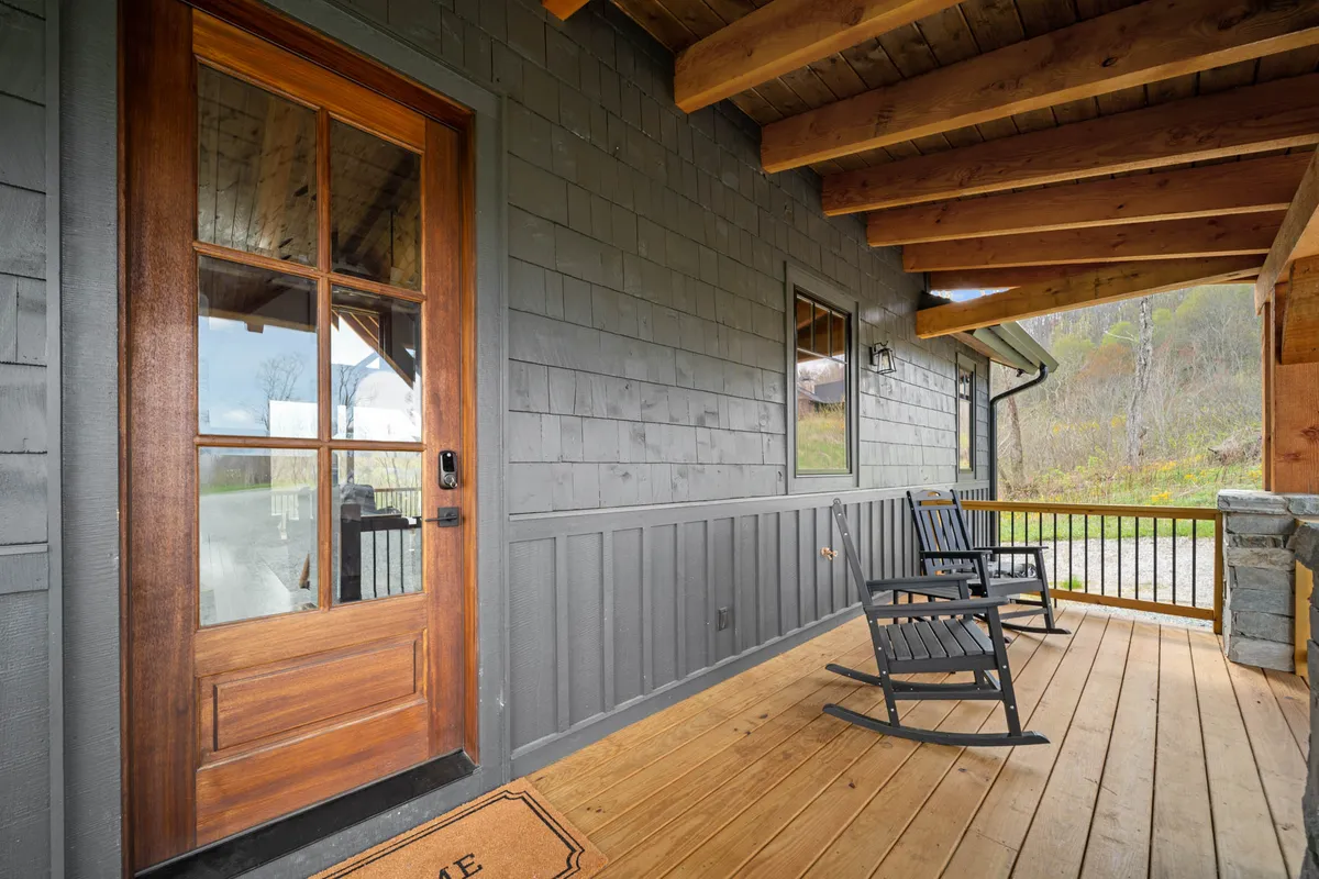 This image shows a welcoming exterior deck area with wooden rocking chairs and a wooden door leading into the home. The deck is surrounded by natural greenery and is an inviting space to relax and enjoy the outdoors, ideal for both people and pets. The atmosphere exudes mountain charm and comfort, hinting at peaceful mornings or lazy afternoons spent outside with furry friends.