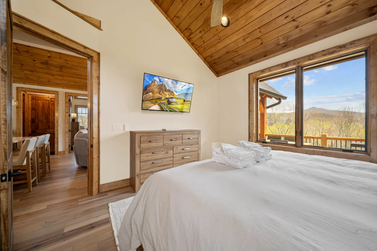 A tranquil bedroom with a king-size bed adorned with soft, neatly arranged towels, a wooden dresser, and expansive windows that frame breathtaking mountain views. The room is bathed in natural light and exudes serenity, inviting relaxation and restful mornings.