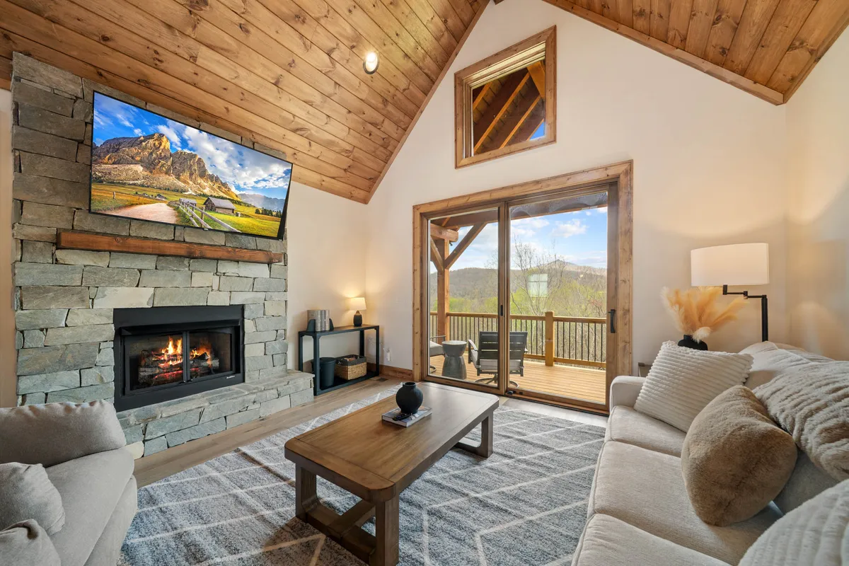 This image captures the core of the great room with its stone fireplace, expansive windows flooding the space with natural light, and comfortable seating arranged for socializing and relaxation. The scene radiates warmth and togetherness, featuring the open layout and stunning mountain views called out in the section.