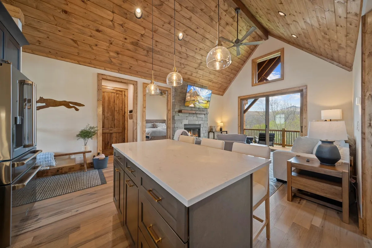 This image offers a wide-angle perspective that includes the kitchen island, stone fireplace, large windows, and wooden ceiling. It demonstrates the seamless connection between cooking, dining, and lounging areas, reinforcing the open-concept layout and flexible use of space.