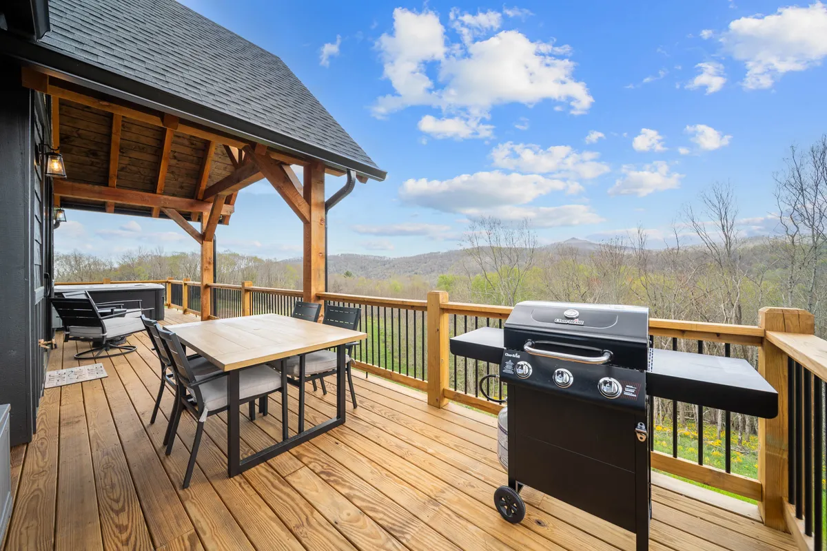 An elevated wooden deck with a dining table, grill, and lounge space overlooking the mountains. The scene is set for outdoor dining, relaxation, and casual gatherings, with expansive views stretching into the horizon.