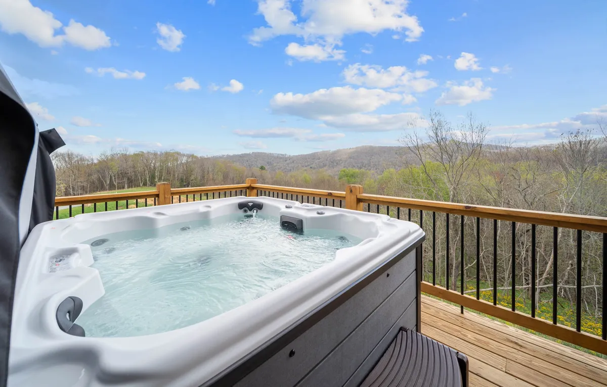 A serene mountain-view deck with a bubbling hot tub in the foreground, framed by a rustic railing. The hot tub invites relaxation with sweeping views of the rolling mountains beyond, while guests can be seen enjoying the tranquil atmosphere or socializing with friends and family.