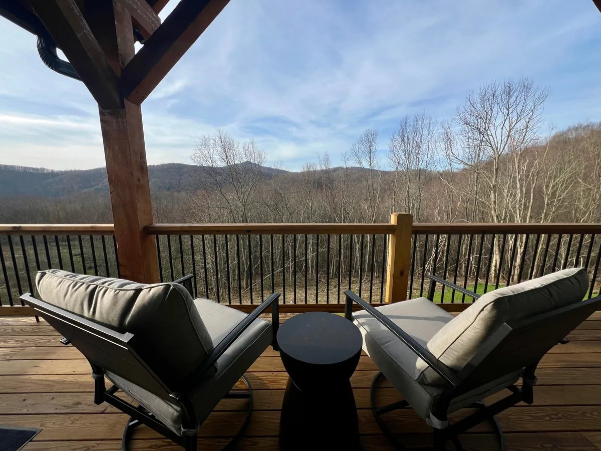 A pair of comfortable chairs and a small circular table are positioned on a deck overlooking a sweeping mountainous landscape. The setting is tranquil, perfect for savoring a morning coffee or simply taking in the stunning natural views with a pet by your side.