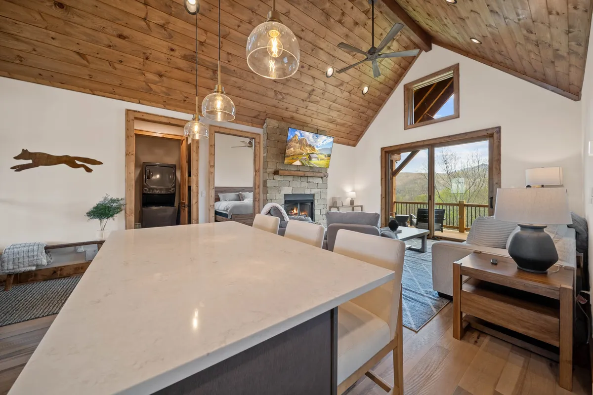 A broader perspective of the kitchen area with a prominent island, adjacent to a cozy fireplace and large windows. The open layout connects the kitchen to the living and dining spaces, with views extending to the scenic deck outside.
