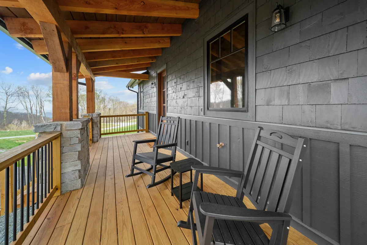 A pair of classic rocking chairs sit on a spacious covered porch, flanked by wooden beams and stone pillars, overlooking a natural landscape. The setting is serene, designed for sipping morning coffee or watching sunsets in comfort.