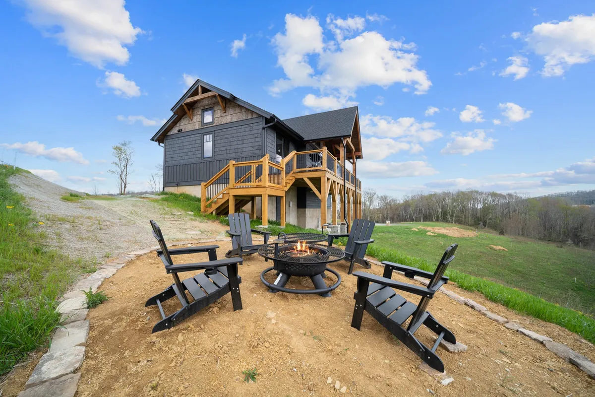 The image showcases the exterior of a newly built single-family house nestled in a serene, wooded setting. At the center is a cozy fire pit surrounded by inviting Adirondack chairs, perfect for gatherings. The spacious deck overlooks beautiful natural scenery, hinting at opportunities for barbecuing, relaxing, and taking in sunset views. The tranquil ambiance is complemented by an aura of relaxation and social connection, making it an ideal spot for evening stargazing or enjoying the fresh mountain air.