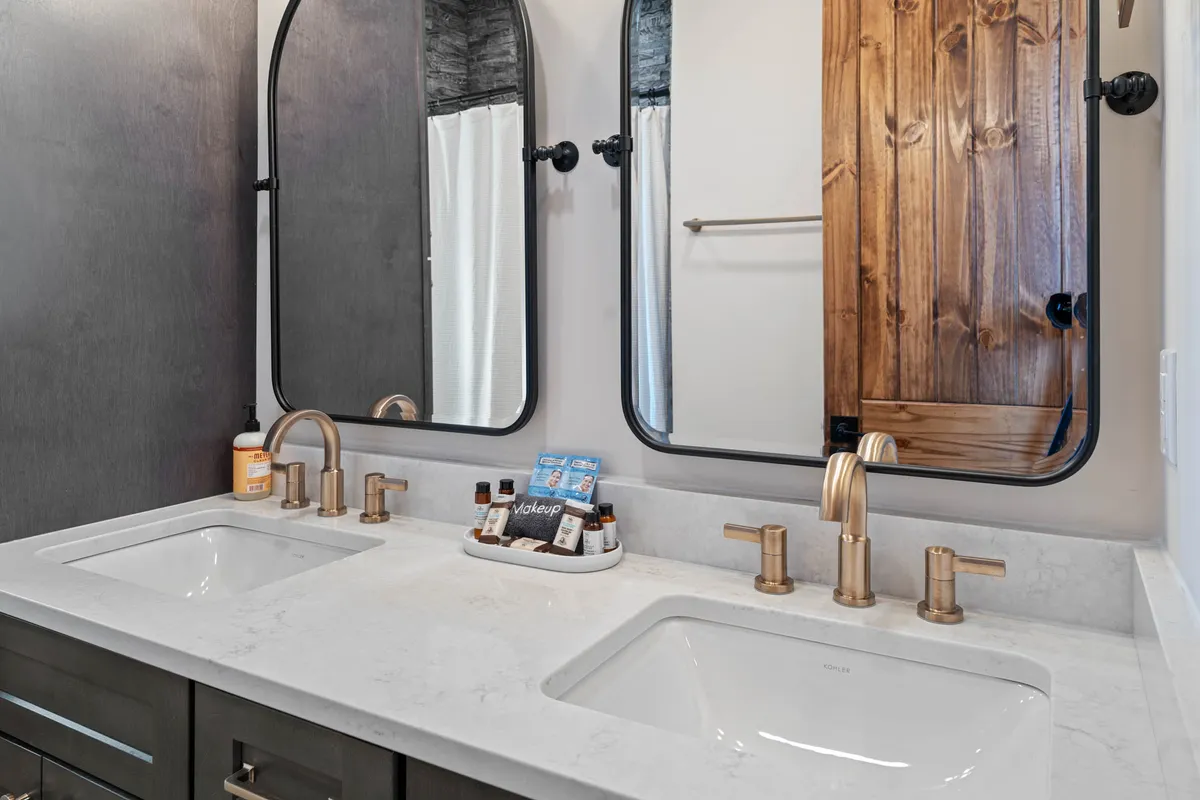 A spacious ensuite bathroom with dual sinks set in a wooden vanity, large mirrors, and a wooden door. The calming, inviting decor suggests a spa-like environment ideal for couples or solo self-care.
