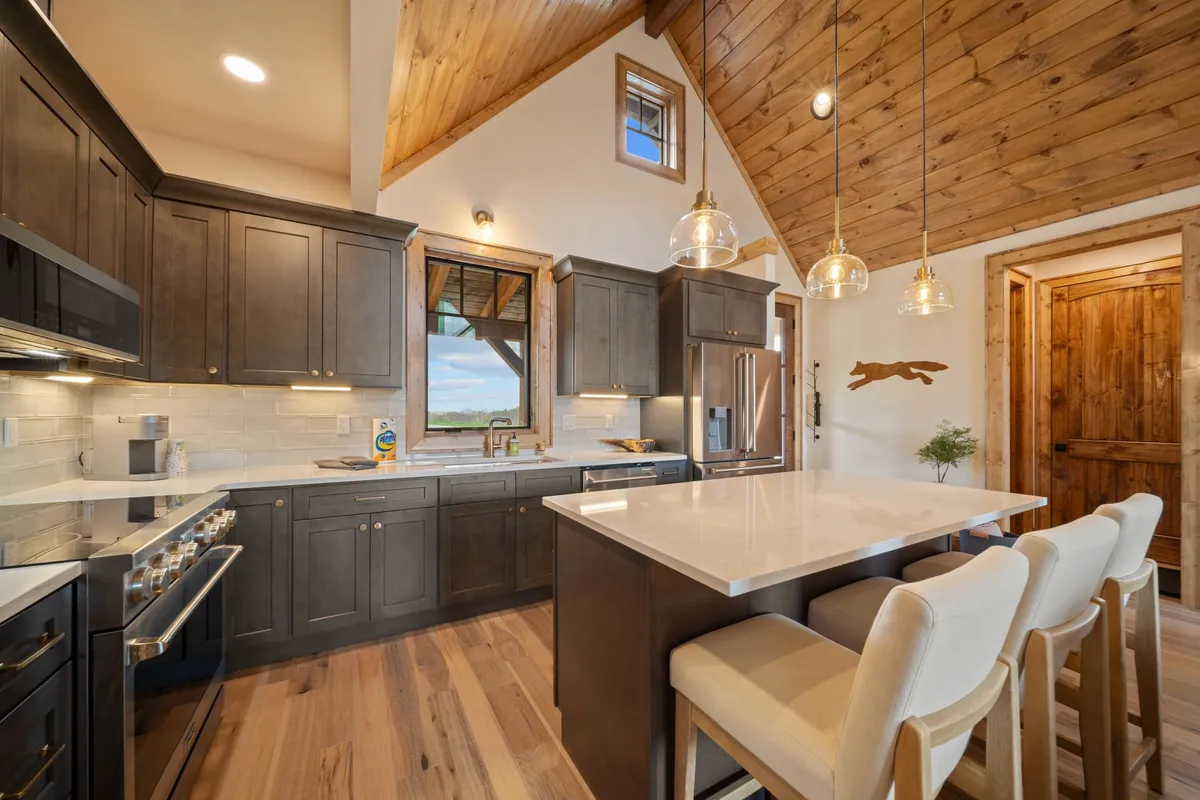 A warm, inviting kitchen scene featuring a prominent island countertop, custom cabinetry, hanging light fixtures, and a wooden ceiling. The island is spacious and ready for meal prep, while natural light pours in from a nearby window. The open layout hints at seamless entertaining and family meals.